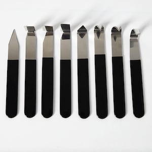 May include: A set of eight metal sculpting tools arranged in a row. Each tool features a long, black, rectangular handle and a distinct silver-coloured metal tip, including pointed, flat, hooked, and curved shapes for precision crafting work.