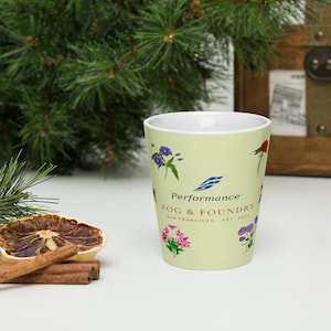 May include: A light green ceramic mug featuring a white interior and a tapered shape. The exterior displays colorful floral illustrations and the text Performance FOG & FOUNDRY SAN FRANCISCO EST. 2028 printed in dark brown and blue lettering.