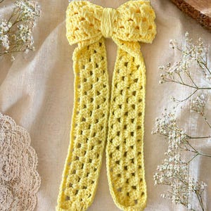 May include: A handmade, yellow crochet bow with long, openwork tails. The bow is made of soft yarn and has a textured appearance. The bow is a hair accessory, perfect for adding a touch of handmade charm to any hairstyle.
