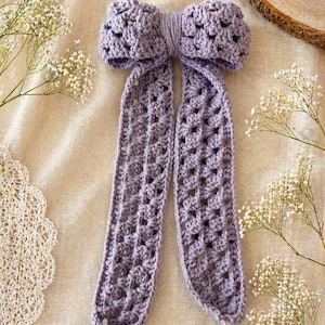 May include: A lavender crochet bow with long, openwork tails. The bow is centered and surrounded by sprigs of white flowers on a neutral background. The bow is handmade and has a textured appearance.