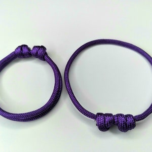 May include: Two purple friendship bracelets with knotted ends. The bracelet on the left has a simple knot, while the bracelet on the right has a more elaborate knot.