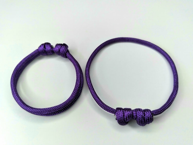 May include: Two purple friendship bracelets with knotted ends. The bracelet on the left has a simple knot, while the bracelet on the right has a more elaborate knot.