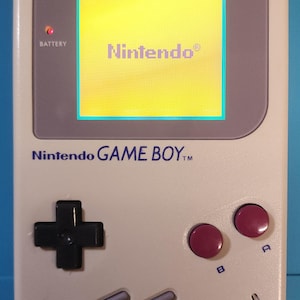 Op de afbeelding: Een lichtgrijze Nintendo Game Boy handheld console. Het scherm toont het Nintendo-logo. Voorzien van een D-pad, A- en B-knoppen en selecteer- en startknoppen. De tekst "Nintendo GAME BOY" is zichtbaar.