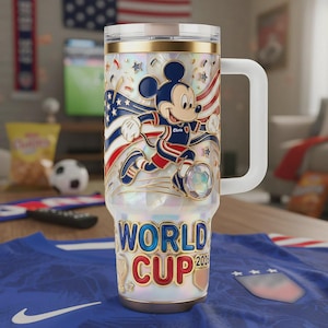 Op de afbeelding: Een witgouden ge&iuml;soleerde beker met Mickey Mouse in een voetbaluniform, met een Amerikaans vlagontwerp. De beker heeft de woorden "World Cup 2026" en een witte handgreep. Een voetbal en een blauw shirt staan op de achtergrond.