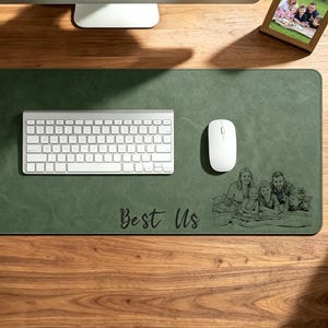 Op de afbeelding: Een groene bureaumat met een wit toetsenbord, muis en de tekst "Best Us". Een familiefoto staat in de rechter benedenhoek. Een computermonitor en een kleine potplant zijn ook zichtbaar.