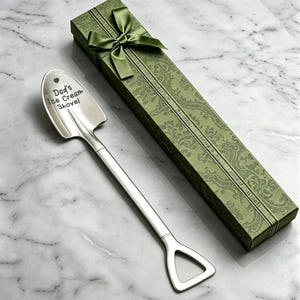 Op de afbeelding: Een zilveren ijsschep met de tekst "Dad's Ice Cream Shovel" gegraveerd op de schep. Gepresenteerd met een groene geschenkdoos en een bijpassende strik. De doos heeft een decoratief patroon.