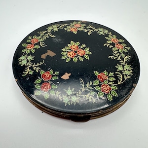 May include: A round black metal compact case featuring a floral pattern of red roses and green leaves arranged in a circular wreath design with a central cluster of roses. The surface shows signs of wear with small areas of paint loss.