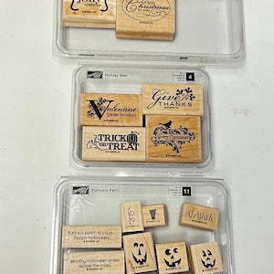 May include: Three clear plastic cases containing sets of wooden rubber stamps. The stamps feature various holiday-themed designs and text, including Christmas, Halloween, and Thanksgiving motifs, with Stampin' Up branding on each wooden block.