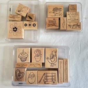 May include: Three clear plastic trays containing various wooden rubber stamps. The stamps feature black ink illustrations of flowers, a cupcake, a duck, an egg, a wagon, and hearts, along with text phrases like "Your friendship makes my heart happy."