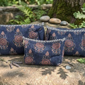 May include: Three quilted fabric pouches in varying sizes, featuring a dark blue base with a repeating floral pattern in rust orange and black. Each pouch has a zippered top closure accented with a black and white striped fabric trim.