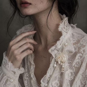 May include: A white lace blouse featuring a high ruffled collar and long sleeves with pearl buttons. A decorative brooch shaped like a shell with dangling white pearls is pinned to the front of the garment, which is crafted from intricate floral lace.
