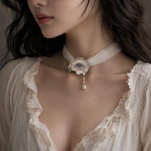 May include: A cream-colored fabric choker necklace featuring a central sculpted eye ornament with a blue iris. The piece is embellished with small white pearls and includes a delicate dangling chain with a single pearl drop at the bottom end.