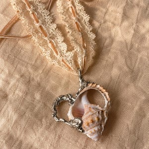 Pode incluir: Um colar com um pingente em forma de coração feito de uma concha natural parcialmente inserida numa moldura de metal prateado texturizado. O pingente está suspenso numa fita de renda bege com uma faixa central lisa e acetinada.