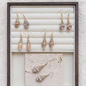 Puede incluir: Cinco pares de pendientes colgantes con caracoles en espiral naturales sujetos a ganchos de metal dorado. Las conchas varían en tonos bronceado, marrón y blanco, algunas con detalles metálicos dorados envueltos alrededor de la superficie.