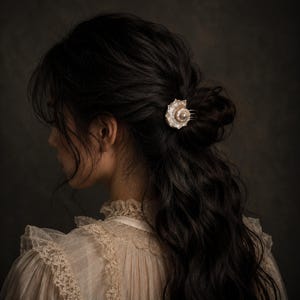 Könnte beinhalten: Ein Haarkamm mit einem kreisförmigen, irisierenden Perlmutt-Ornament, das in der Mitte mit einer kleinen, runden weißen Perle besetzt ist. Das Dekorationselement ist an einem Metallkamm befestigt, der eine Partie dunkles, welliges Haar hält.