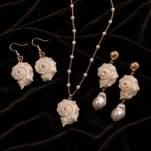 Puede incluir: Un conjunto de joyería que incluye un collar de cadena dorada con pequeñas perlas blancas y un colgante de concha en espiral, un par de pendientes de concha y un par de pendientes con una bola dorada, una concha en espiral y una perla.