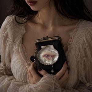 May include: A black leather kiss-lock coin purse featuring a decorative seashell appliqué on the front. The shell contains a realistic eye illustration and is embellished with small white pearls and a single dangling pearl charm on a silver chain.