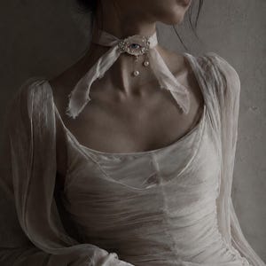 May include: A white fabric choker featuring a central oval ornament with a realistic eye illustration, surrounded by small white beads. Two delicate silver chains hang from the ornament, each ending in a single white pearl drop. The choker is tied.