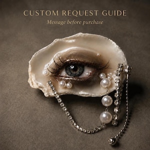 Puede incluir: Una concha de ostra natural con un ojo humano pintado con pestañas largas, adornado con pequeñas perlas blancas y una cadena plateada colgante con pedrería transparente. El texto dice: CUSTOM REQUEST GUIDE Message before purchase.