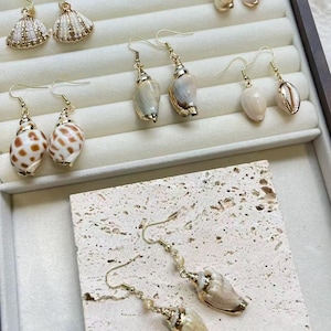 Peut inclure: Une collection de boucles d'oreilles à crochet dorées ornées de divers coquillages naturels. L'ensemble comprend des coquillages en éventail, des cauris tachetés et des coquillages en spirale, suspendus à des crochets dorés et des perles.