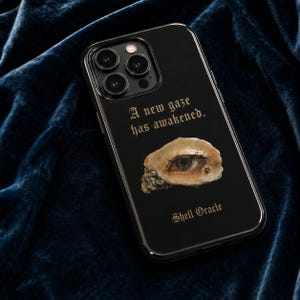 Puede incluir: Una funda de smartphone negra con el texto A new gaze has awakened y Shell Oracle en una fuente gótica. El centro de la funda muestra un gráfico realista de un ojo humano incrustado en un fragmento de concha de color beige texturizado.