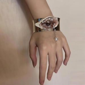 May include: A wide silver-toned metal cuff bracelet featuring a central sculptural element resembling an eye encased in a shell-like structure. The design is embellished with small white pearls and includes a delicate chain drop with a single pearl.