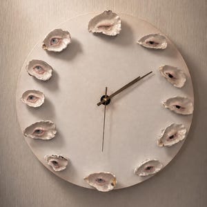 Puede incluir: Un reloj de pared circular con una esfera de color blanco roto mate y manecillas finas de tono dorado. Doce conchas de ostra individuales están dispuestas alrededor del perímetro, cada una con un ojo humano pintado de forma realista.