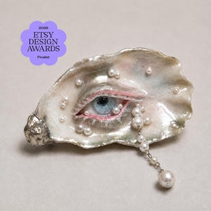 May include: An oyster shell sculpture featuring a realistic blue eye with pink eyelids and white eyelashes, decorated with scattered white pearls and a dangling pearl chain. A purple badge reads 2026 ETSY DESIGN AWARDS Finalist.