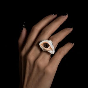 May include: A ring featuring a realistic sculpted human eye with a brown iris and pale skin-toned eyelids. The eye is set within a textured, irregular white base that wraps around the finger, creating a detailed and lifelike appearance.
