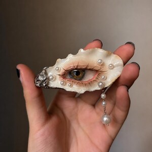 Può includere: Una spilla decorativa realizzata con un guscio di ostrica naturale che presenta un occhio dipinto in modo realistico con ciglia lunghe. Il guscio è decorato con diverse piccole perle bianche e una catenella argentata con tre perle.