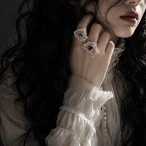May include: Two rings worn on the fingers, each featuring a realistic sculpted eye design. The top ring displays a blue iris, while the bottom ring features a brown iris. Both rings have irregular, textured, pale-toned settings surrounding the eyes.