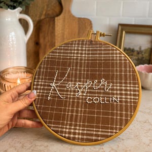 May include: A circular embroidery hoop containing brown and white plaid fabric. The name Kasper is embroidered in white cursive thread across the centre, with the name COLLIN embroidered in white capital letters directly underneath the name Kasper.