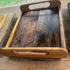 May include: A rectangular wooden serving tray with a dark stained interior and natural wood frame. The tray has two handles with oval cutouts. The wood grain is visible throughout the tray.