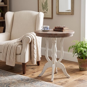 May include: A round side table with a natural wood-toned top and a white, distressed-finish pedestal base featuring four curved legs. The table surface holds a blue and white patterned teacup on a saucer and two stacked brown books with glasses.