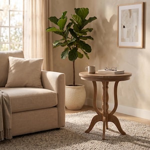 May include: A round wooden side table with a pedestal base featuring curved legs. On the tabletop sits a two-toned ceramic mug, a closed book, and a pair of reading glasses. The table is positioned next to a beige upholstered sofa and a potted plant.