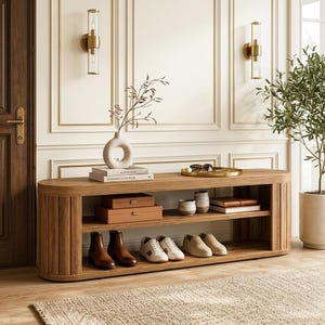 May include: A long, oval-shaped wooden entryway bench with two open shelves. The top surface holds a white donut-shaped vase, two stacked books, and a gold tray with sunglasses. The shelves contain two tan boxes, two ceramic mugs, books, and three pairs of shoes.