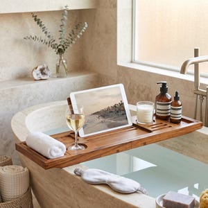 May include: A rectangular wooden bathtub tray featuring a built-in stand for a tablet, a circular cutout for a wine glass, and a recessed area for a candle. The tray holds a white rolled towel, a glass of white wine, and two amber bottles.