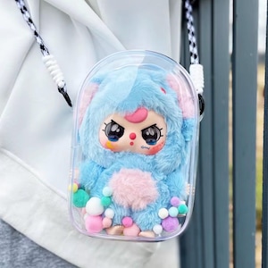 May include: A clear, plastic crossbody bag featuring a plush, blue monster character with large eyes and a pink nose. The character has a fluffy blue body and a pink belly. The bag has a black and white strap and is decorated with colorful pom-poms.