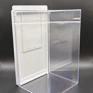 K&ouml;nnte beinhalten: Eine durchsichtige Acryl-Vitrine mit einer wei&szlig;en R&uuml;ckwand, die eine vertikal gerippte Textur und einen rechteckigen Einsatz aufweist. Die Oberkante der wei&szlig;en Platte ist mit dem Text GARAGE KIT in Gro&szlig;buchstaben gepr&auml;gt.