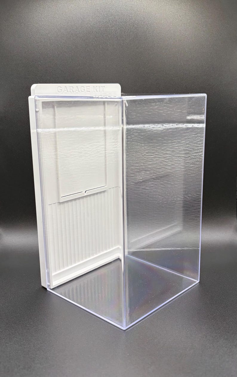 K&ouml;nnte beinhalten: Eine durchsichtige Acryl-Vitrine mit einer wei&szlig;en R&uuml;ckwand, die eine vertikal gerippte Textur und einen rechteckigen Einsatz aufweist. Die Oberkante der wei&szlig;en Platte ist mit dem Text GARAGE KIT in Gro&szlig;buchstaben gepr&auml;gt.
