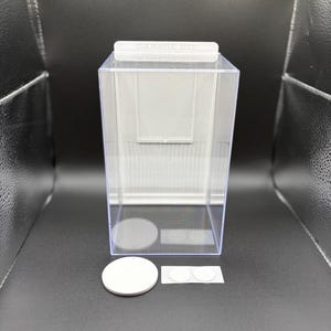 K&ouml;nnte beinhalten: Eine durchsichtige, rechteckige Vitrine mit wei&szlig;er R&uuml;ckwand und einer oberen Lasche mit der Aufschrift GARAGE KIT. Enthalten sind ein gro&szlig;er wei&szlig;er runder Sockel sowie zwei kleinere wei&szlig;e, runde Klebepunkte zur Befestigung von Objekten.