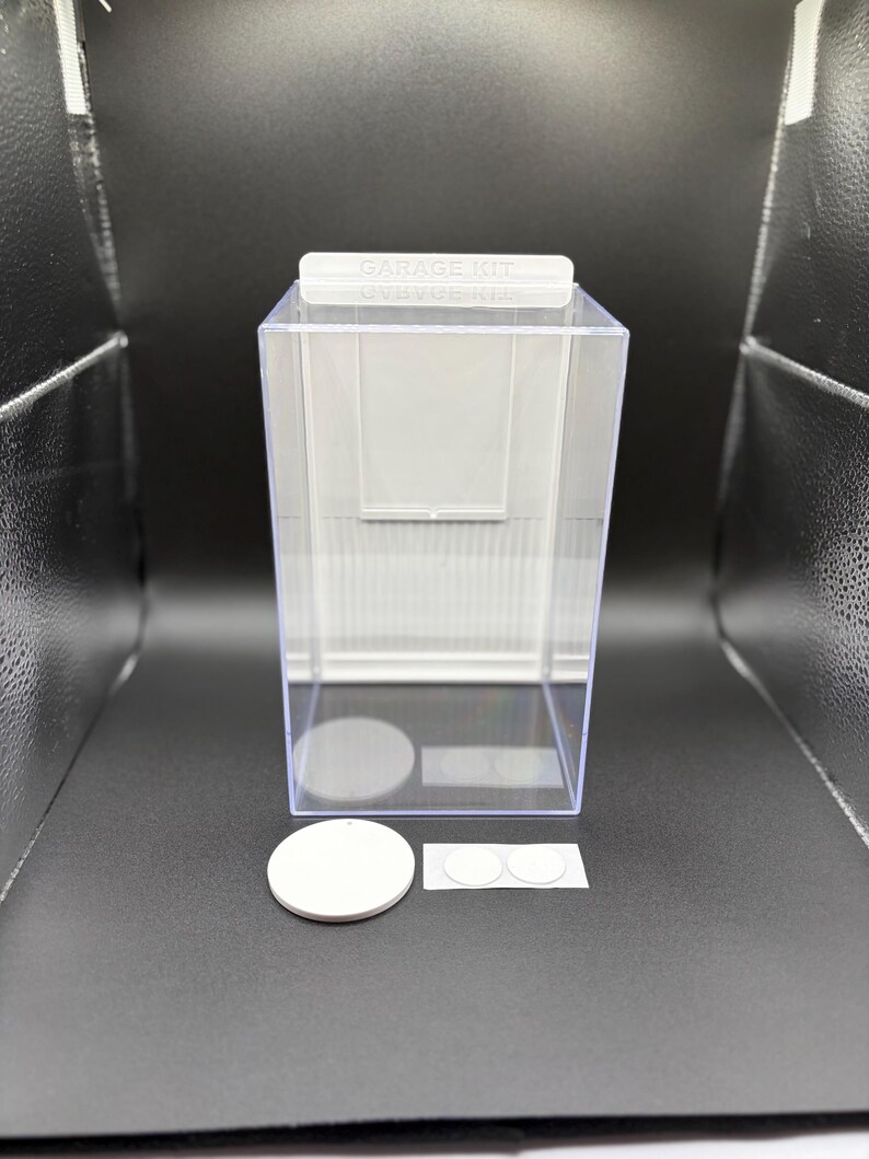 K&ouml;nnte beinhalten: Eine durchsichtige, rechteckige Vitrine mit wei&szlig;er R&uuml;ckwand und einer oberen Lasche mit der Aufschrift GARAGE KIT. Enthalten sind ein gro&szlig;er wei&szlig;er runder Sockel sowie zwei kleinere wei&szlig;e, runde Klebepunkte zur Befestigung von Objekten.