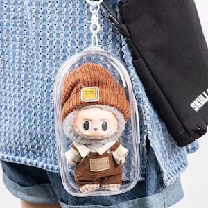 May include: A small doll wearing a brown knit cap with a yellow patch, a gray fur-lined collar, and brown overalls. The doll is encased in a clear plastic case attached to a keychain. A black bag with the word "SKULL" is visible in the background.