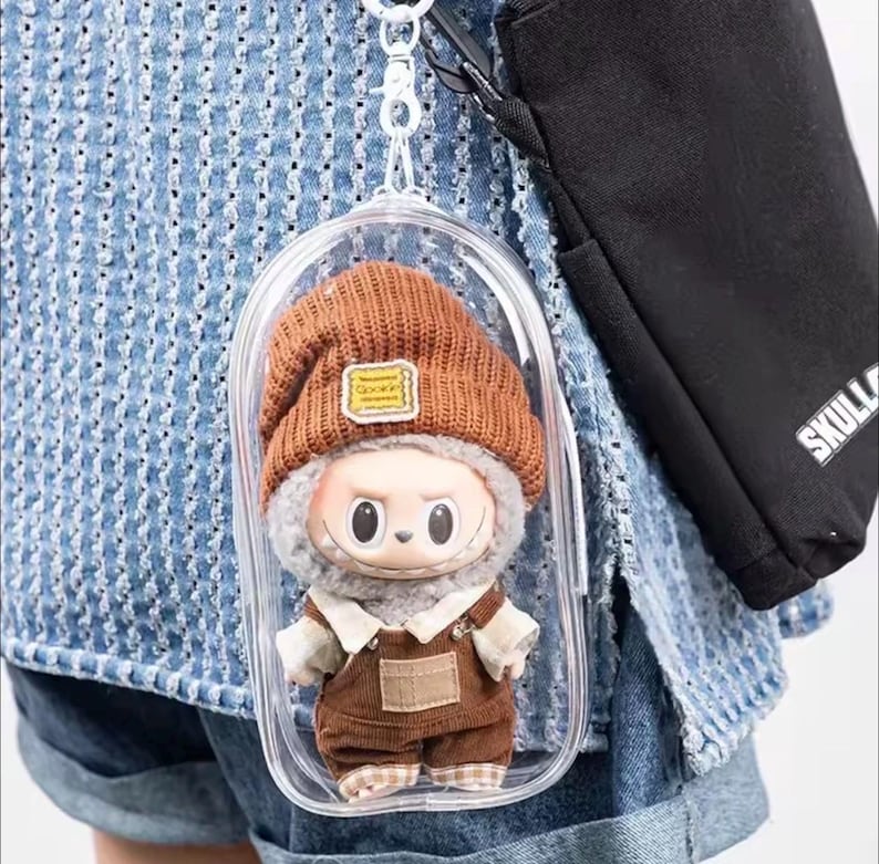 May include: A small doll wearing a brown knit cap with a yellow patch, a gray fur-lined collar, and brown overalls. The doll is encased in a clear plastic case attached to a keychain. A black bag with the word "SKULL" is visible in the background.