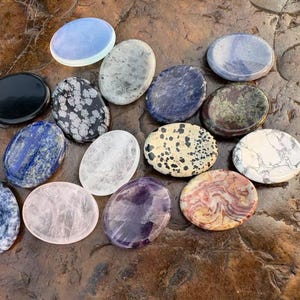 Puede incluir: Una colección de piedras preciosas ovaladas en varios colores y patrones. Las piedras incluyen variedades negras, azules, blancas, rosas y estampadas. Las piedras preciosas se exhiben sobre una superficie marrón texturizada.