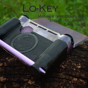 May include: Black and silver Magsafe grip, stand, and stylus holder for AYN Thor, with the text "LO-KEY". The holder has a purple stylus and accents. The product is resting on a dark brown, textured surface.