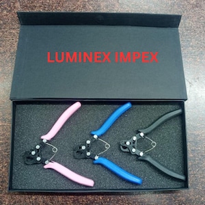 May include: A set of three pliers with pink, blue, and black handles, displayed in a black box. The text "LUMINEX IMPEX" is printed in red on the inside of the box lid. The pliers are designed for a specific task.
