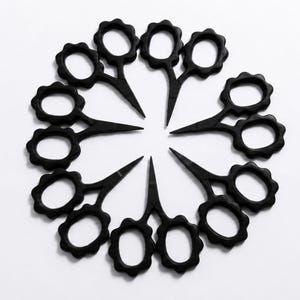 Può includere: Una collezione di dieci piccole forbici nere disposte in un motivo circolare. Ogni paio di forbici presenta un caratteristico design dell'impugnatura smerlato o a forma di fiore con lame appuntite, tutte orientate verso il centro del cerchio.
