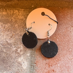 May include: A pair of black fabric circle earrings with silver-colored hooks. The earrings are displayed on a white tag with a black string. The background is a textured, reddish-brown surface.
