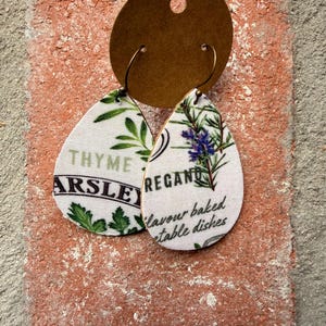 May include: Teardrop earrings with a white background, featuring green herb illustrations and text. One earring says "Thyme Parsley," and the other says "Oregano flavour baked vegetable dishes." The earrings hang from gold-coloured hooks and a brown tag.